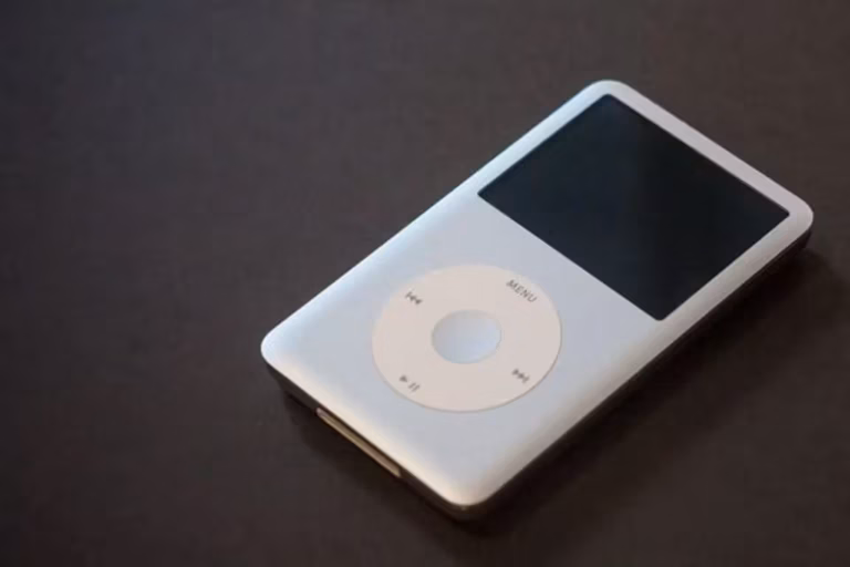 Un iPod Classic argenté, doté d'un écran noir et d'une molette circulaire blanche, repose sur une surface sombre. Le design épuré et minimaliste de l'appareil permet d'accéder facilement aux fichiers, tandis que ses boutons apparents soulignent sa simplicité d'utilisation et son style classique.