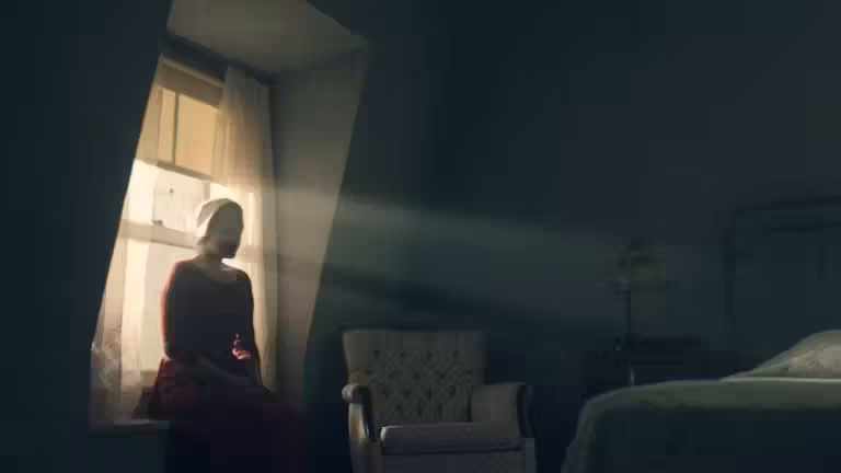 Offred sitting in her room beside a partly open window in The Handmaid’s Tale