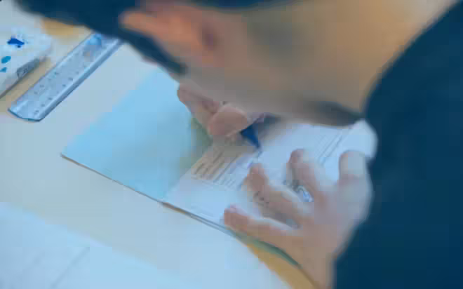 Une personne remplit un formulaire avec un stylo bleu sur un dossier ou un bloc notes bleu clair, travaillant peut être sur un essai en français. L'individu porte une chemise sombre. Une règle et une gomme sont visibles sur la table à côté du formulaire. L'accent est mis sur la main et la forme de la personne, le visage étant partiellement flou et flou.