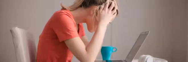 Une personne portant un t shirt orange est assise, penchée sur un ordinateur portable, les mains tenant sa tête en signe de frustration, probablement après avoir reçu des courriels de l'Éducation Nationale dont elle souhaite se désabonner. Une tasse turquoise et des écouteurs blancs sont posés sur la table à côté d'eux, sur un fond uni et clair.