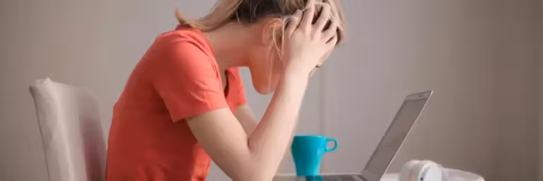 Une personne portant un t shirt orange est assise, penchée sur un ordinateur portable, les mains tenant sa tête en signe de frustration, probablement après avoir reçu des courriels de l'Éducation Nationale dont elle souhaite se désabonner. Une tasse turquoise et des écouteurs blancs sont posés sur la table à côté d'eux, sur un fond uni et clair.
