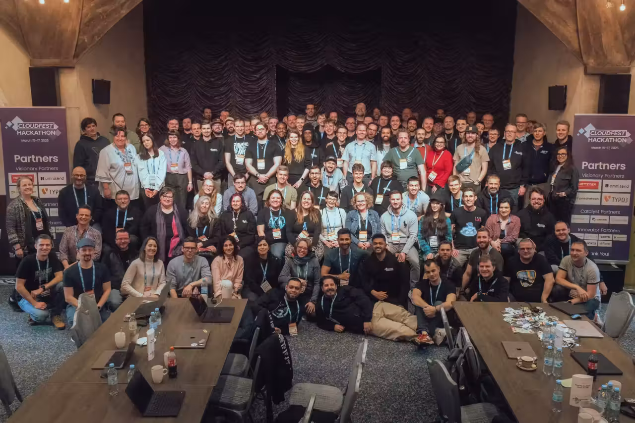 Un grand groupe de personnes, d'âges et de sexes divers, pose et sourit ensemble dans une salle de conférence ornée de poutres en bois et de banderoles sur lesquelles est écrit « CloudFest Hackathon 2025 ». Plusieurs tables avec ordinateurs portables et badges agrémentent ce résumé dynamique du hackathon.