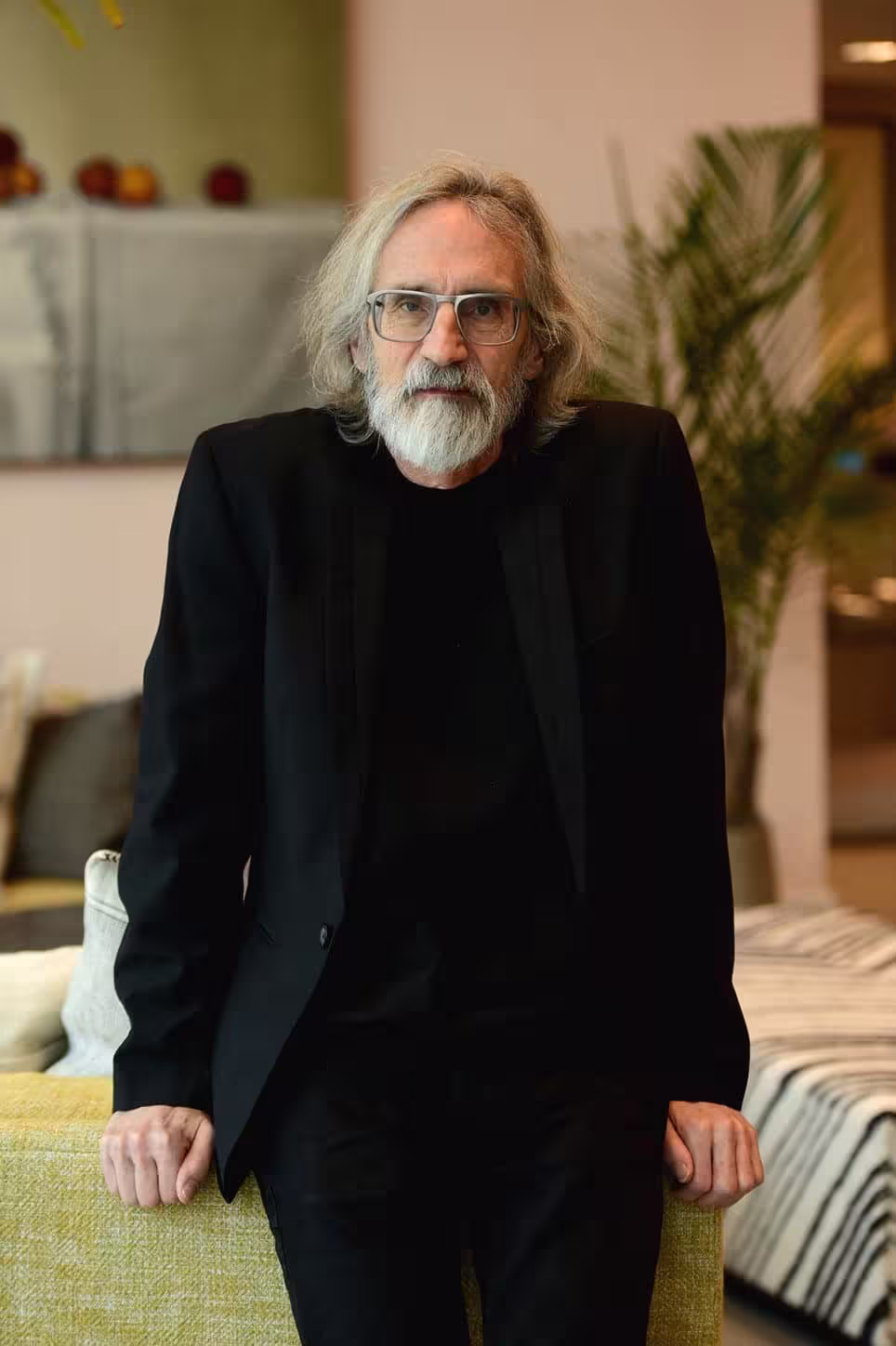 Un homme âgé aux longs cheveux gris, à la barbe blanche et aux lunettes porte un costume et une chemise noirs. Il est adossé à un canapé jaunâtre dans une pièce moderne dont le décor évoque subtilement l'imaginaire médiéval des illustrations de Tolkien de John Howe.