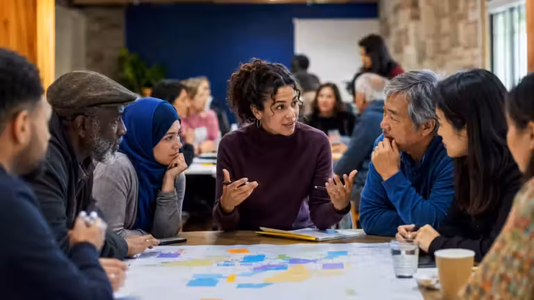 Six personnes sont assises autour d'une table recouverte de cartes et de documents colorés, en pleine discussion. Une femme, assise au centre, accompagne ses propos de gestes, soulignant l'importance de la médiation et du plurilinguisme dans leur travail collaboratif. Le cadre semble être celui d'une réunion ou d'un atelier consacré à la communication multilingue, tandis que d'autres groupes discutent en arrière-plan.