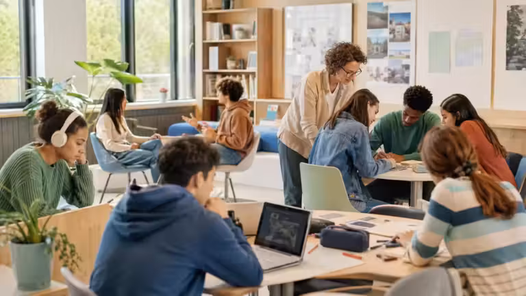 Une salle de classe où les élèves travaillent en groupes autour de tables. Certains apprenants lisent, écrivent ou utilisent des ordinateurs portables, tandis qu’un groupe discute avec un enseignant qui se penche vers eux pour les aider, favorisant ainsi l’autonomie et la différenciation dans leurs activités. La salle est lumineuse, avec de grandes fenêtres, des étagères remplies de livres et des affiches pédagogiques accrochées aux murs.